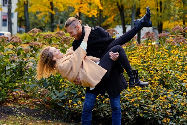 El hombre barbudo pelirrojo sostiene en sus brazos a una linda mujer rubia en un parque de otoño.