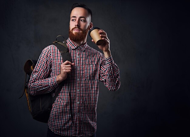Hombre barbudo pelirrojo vestido con una camisa de lana sostiene una mochila y se lleva una taza de café.