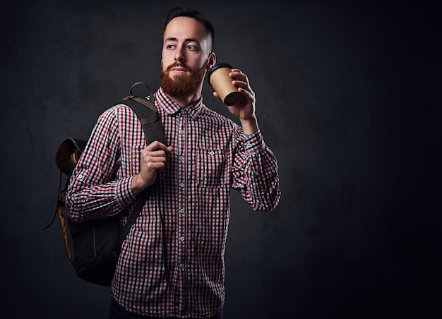 Hombre barbudo pelirrojo vestido con una camisa de lana sostiene una mochila y se lleva una taza de café.