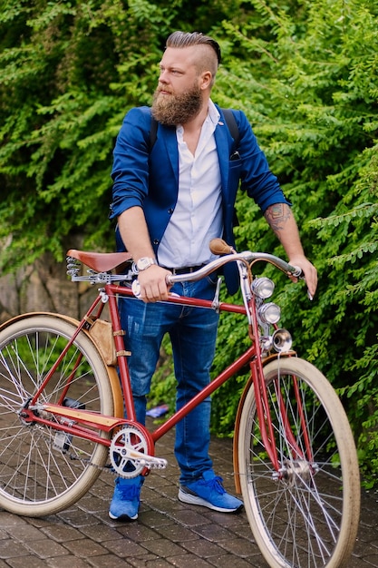 Hombre barbudo pelirrojo vestido con una chaqueta azul y jeans en una bicicleta retro en un parque.