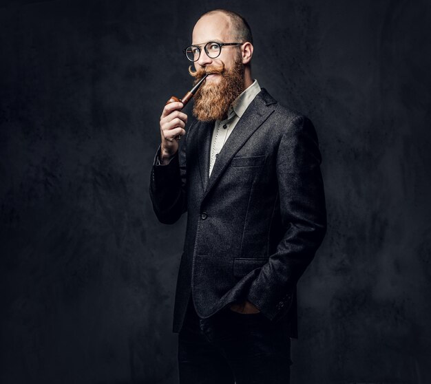 Hombre barbudo pelirrojo vestido con traje y anteojos fumando pipa tradicional sobre fondo gris oscuro.