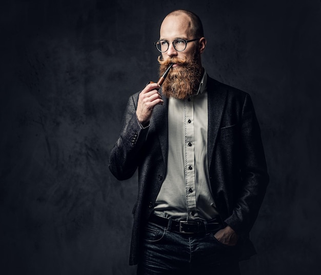 Hombre barbudo pelirrojo vestido con traje y anteojos fumando pipa tradicional sobre fondo gris oscuro.