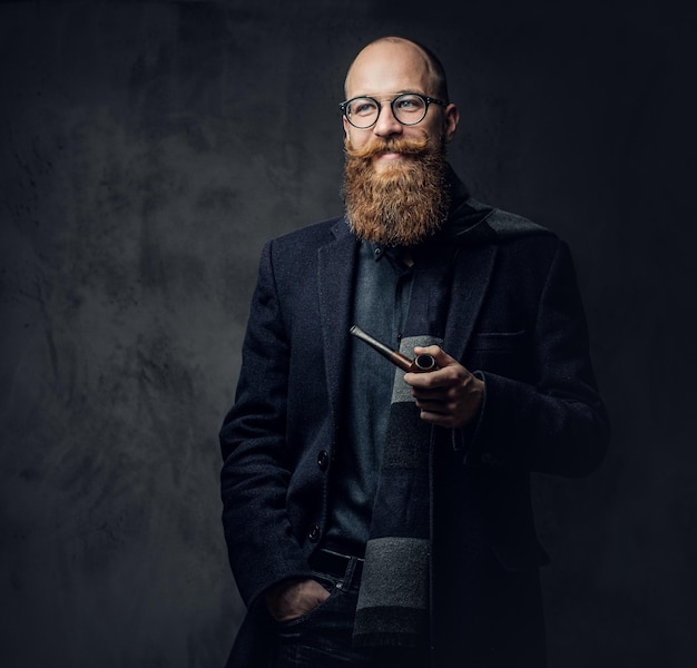 Hombre barbudo pelirrojo vestido con traje y anteojos fumando pipa tradicional sobre fondo gris oscuro.