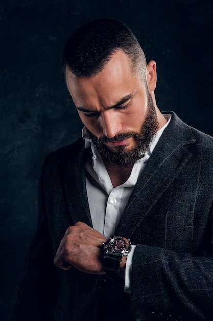 Un hombre barbudo pensativo con traje oscuro está comprobando qué hora es ahora en un estudio fotográfico oscuro.