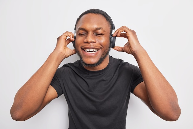 El hombre barbudo de piel oscura positiva disfruta de un sonido perfecto en auriculares estéreo mantiene los ojos cerrados y sonríe ampliamente