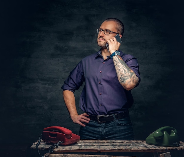 Hombre barbudo positivo en anteojos usando un teléfono inteligente en una habitación con teléfonos celulares analógicos.