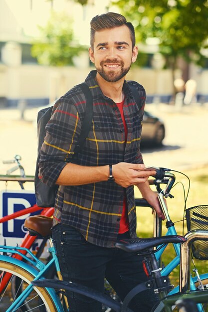 Foto gratuita un hombre barbudo positivo vestido con una camisa de lana usando un teléfono inteligente cerca del estacionamiento de bicicletas.