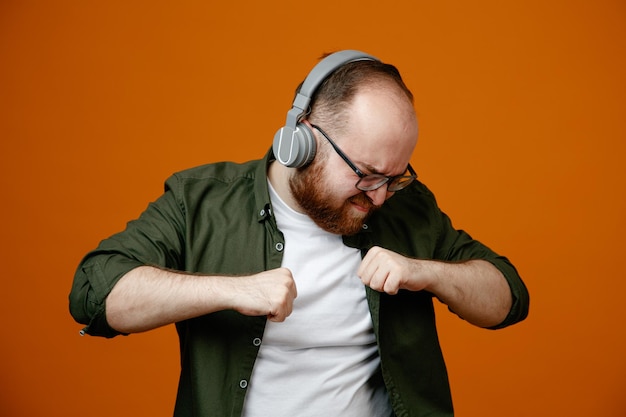 Hombre barbudo en ropa casual con gafas con auriculares feliz y emocionado bailando escuchando música de pie sobre fondo naranja