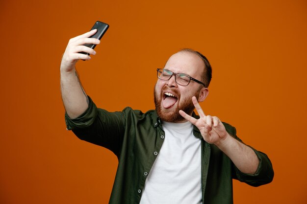 Hombre barbudo en ropa casual con gafas haciendo selfie usando un teléfono inteligente feliz y alegre sacando la lengua mostrando vsign de pie sobre fondo naranja