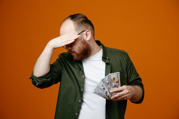 Hombre barbudo en ropa casual con gafas sosteniendo dinero en efectivo mirando estresado cubriendo los ojos con la mano de pie sobre fondo naranja