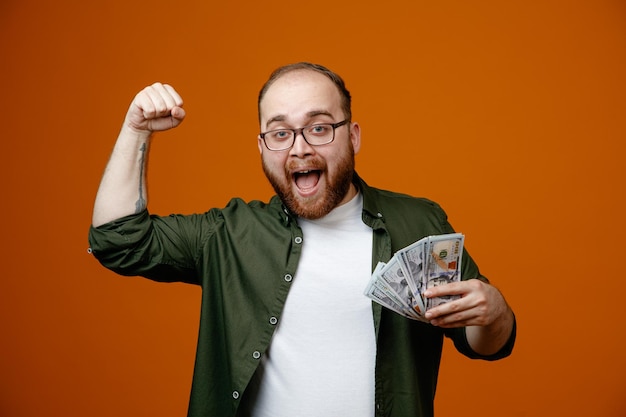 Hombre barbudo en ropa casual con gafas sosteniendo efectivo apretando el puño feliz y positivo regocijándose de su éxito de pie sobre fondo naranja