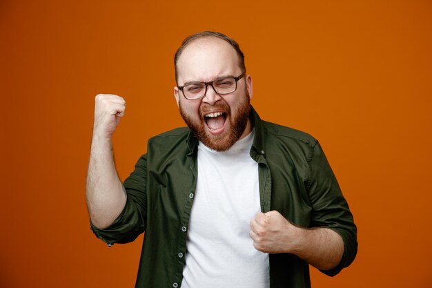 Hombre barbudo con ropa informal apretando los puños loco feliz regocijándose de su éxito de pie sobre fondo naranja