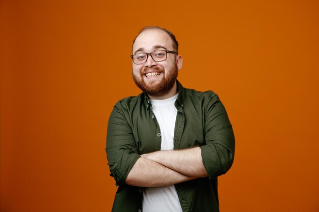 Hombre barbudo con ropa informal cruzando las manos en el pecho sonriendo alegremente feliz y positivo de pie sobre fondo naranja