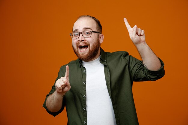 Hombre barbudo con ropa informal y gafas apuntando con el dedo índice hacia arriba feliz y complacido sonriendo alegremente de pie sobre un fondo naranja