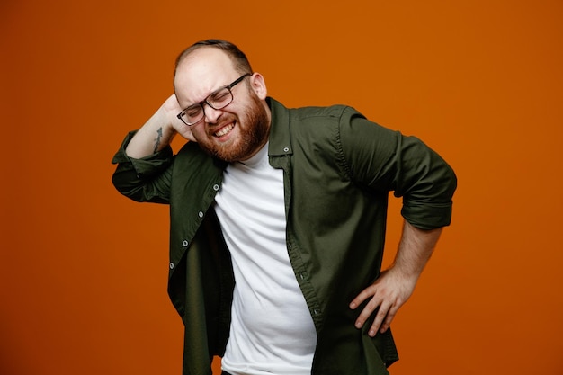 Hombre barbudo con ropa informal y gafas con aspecto de malestar sintiendo dolor en la espalda y el cuello de pie sobre un fondo naranja