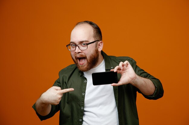 Hombre barbudo con ropa informal y gafas que muestra un teléfono inteligente apuntando con el dedo índice sonriendo alegremente de pie sobre un fondo naranja