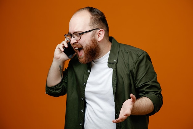 Hombre barbudo con ropa informal y gafas que parece confundido y decepcionado mientras habla por teléfono móvil de pie sobre un fondo naranja