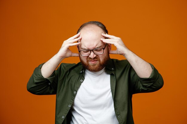 Hombre barbudo con ropa informal y gafas que se ve mal tocándose la cabeza con un fuerte dolor de cabeza parado sobre un fondo naranja