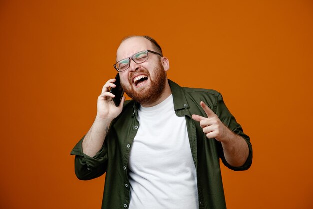 Hombre barbudo con ropa informal y gafas riéndose mientras habla por teléfono móvil de pie sobre un fondo naranja