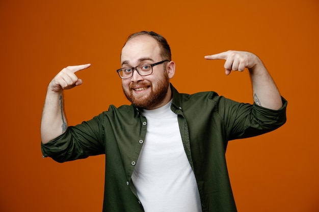 Hombre barbudo con ropa informal y gafas satisfecho de sí mismo apuntándose a sí mismo sonriendo alegremente de pie sobre un fondo naranja