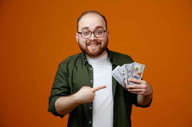 Hombre barbudo con ropa informal y gafas sosteniendo dinero en efectivo apuntando con el dedo índice al dinero feliz y complacido sonriendo de pie sobre un fondo naranja