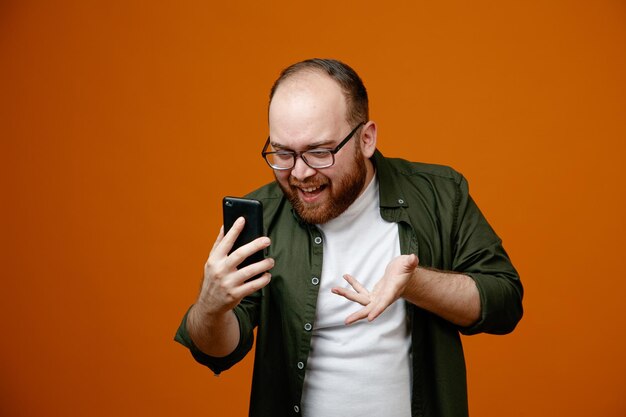 Hombre barbudo con ropa informal con gafas sosteniendo un teléfono inteligente que parece confundido con una videollamada de pie sobre un fondo naranja