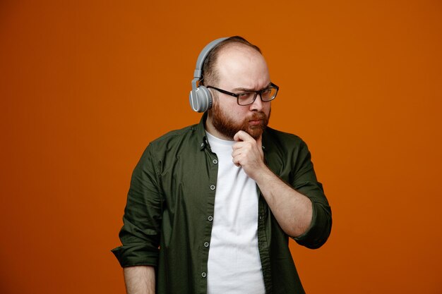 Hombre barbudo con ropa informal que usa anteojos con auriculares mirando a la cámara con expresión pensativa pensando sosteniendo la mano en la barbilla de pie sobre fondo naranja