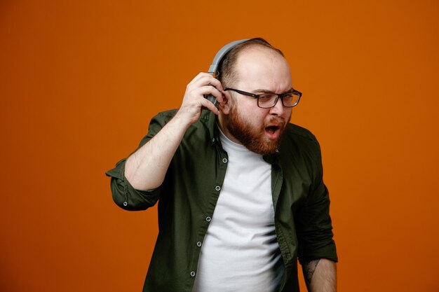 Hombre barbudo con ropa informal usando anteojos con auriculares mirando a la cámara confundido tratando de escuchar mejor quitándose los auriculares de pie sobre fondo naranja