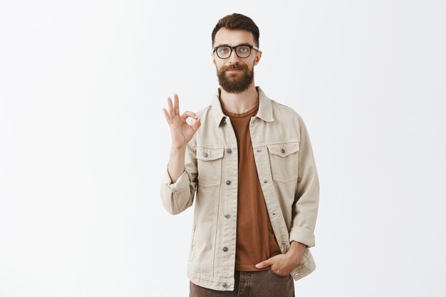 Hombre barbudo satisfecho con gafas posando contra la pared blanca
