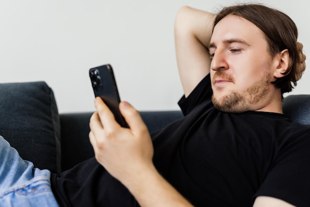 Hombre barbudo seguro está sentado en el sofá y escribiendo en el teléfono inteligente