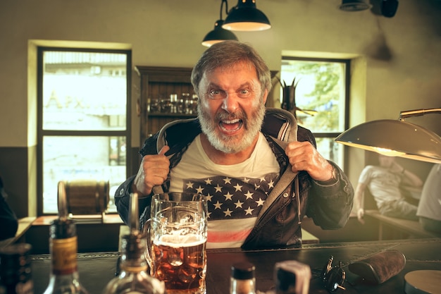 Hombre barbudo senior bebiendo alcohol en un pub y viendo un programa deportivo en la televisión. Disfrutando de mi cerveza y mi cerveza favorita. Hombre con jarra de cerveza sentado a la mesa. Aficionado al fútbol o al deporte. Concepto de emociones humanas