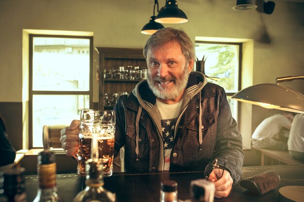 Hombre barbudo senior bebiendo alcohol en un pub y viendo un programa deportivo en la televisión. Disfrutando de mi cerveza y mi cerveza favorita. Hombre con jarra de cerveza sentado a la mesa. Aficionado al fútbol o al deporte. Concepto de emociones humanas