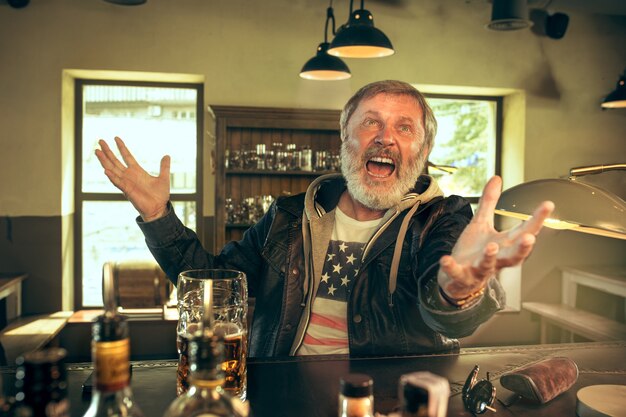 Hombre barbudo Senior bebiendo alcohol en un pub y viendo un programa deportivo en la televisión. Disfrutando de mi cerveza y mi cerveza favorita. Hombre con jarra de cerveza sentado a la mesa. Aficionado al fútbol o al deporte. Concepto de emociones humanas