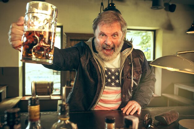 Hombre barbudo senior bebiendo alcohol en un pub y viendo un programa deportivo en la televisión. Disfrutando de mi cerveza y mi cerveza favorita. Hombre con jarra de cerveza sentado a la mesa.