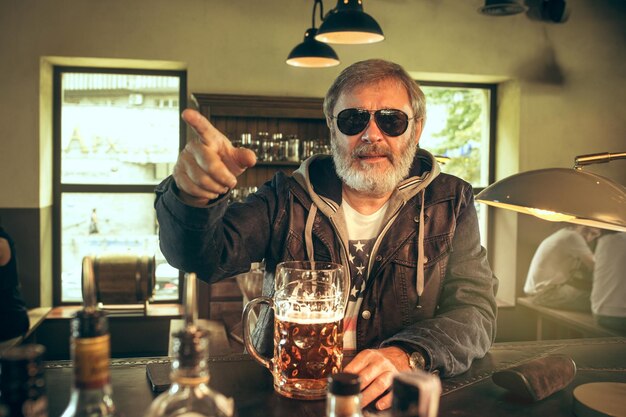 Hombre barbudo senior bebiendo alcohol en un pub y viendo un programa deportivo en la televisión. Disfrutando de mi cerveza y mi cerveza favorita. Hombre con jarra de cerveza sentado a la mesa.