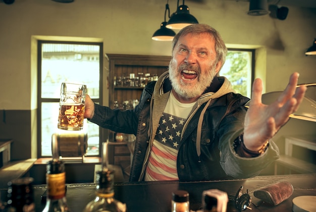 Hombre barbudo senior bebiendo alcohol en un pub y viendo un programa deportivo en la televisión.