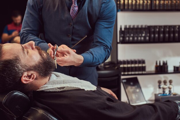 Hombre barbudo sentado en un sillón en una barbería mientras peluquero modela barba con tijeras y peine en la barbería.