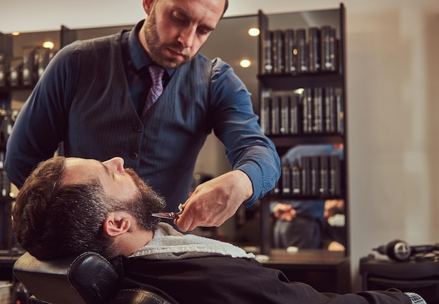 Hombre barbudo sentado en un sillón en una barbería mientras peluquero modela barba con tijeras y peine en la barbería.