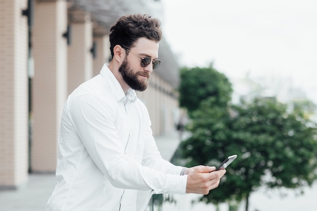Un hombre barbudo, serio y elegante con camisa blanca y gafas de sol de pie en las calles de la ciudad y surfing smartphone