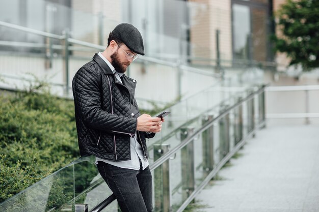 Un hombre barbudo, serio y elegante de pie en las calles de la ciudad cerca del moderno centro de oficinas con teléfono móvil