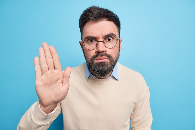 Hombre barbudo serio mantiene la palma levantada hace gesto de restricción o negación usa gafas transparentes redondas puente casual pide que se detenga