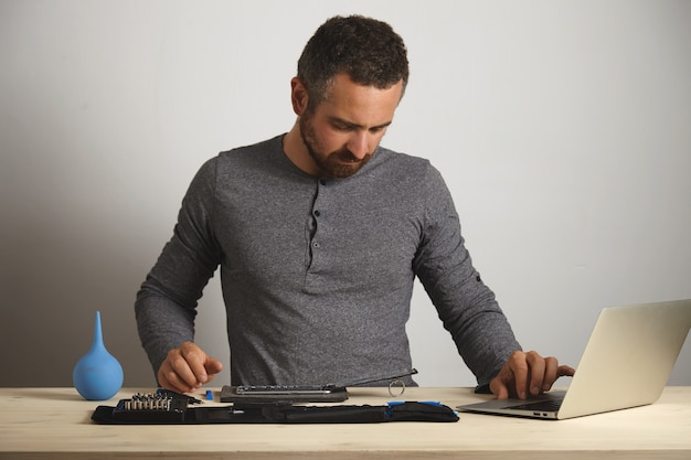 Hombre barbudo serio mirando el teléfono desmontado y trabajando en una computadora portátil para pedir las piezas necesarias para cambiarlo