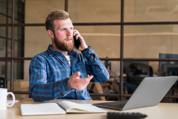 Hombre barbudo serio que habla en el teléfono celular en el lugar de trabajo