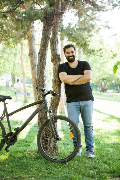 Hombre barbudo sonriente apoyado en un árbol en el parque al lado de su bicicleta