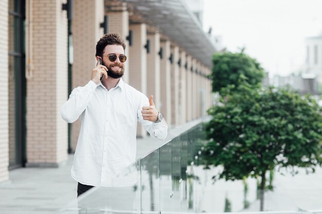 Un hombre barbudo, sonriente y elegante con camisa blanca y gafas de sol de pie en las calles de la ciudad y llamando por teléfono móvil