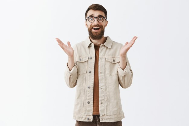Hombre barbudo sorprendido y agradecido con gafas posando contra la pared blanca