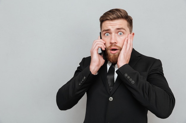 Hombre barbudo sorprendido hablando por su teléfono inteligente con la boca abierta