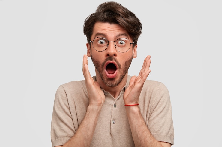 Foto gratuito hombre barbudo sorprendido recibe noticias inesperadas de un amigo, toma las manos cerca de la cara, abre la boca ampliamente, expresa sorpresa, aislado en una pared blanca