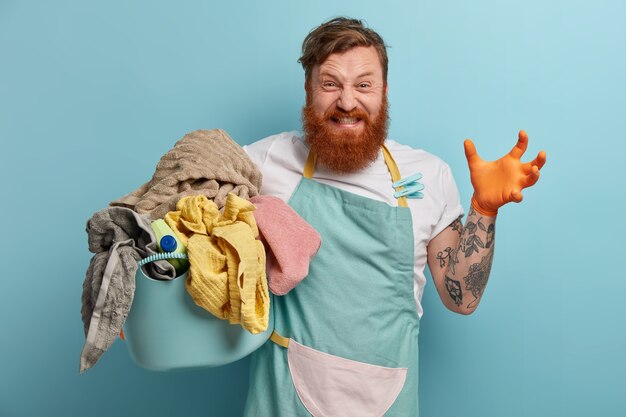 Hombre barbudo sostiene canasta de lavandería, abrumado por las tareas del hogar