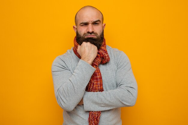 Hombre barbudo en sudadera gris con bufanda alrededor de su cuello mirando con la mano en la barbilla pensando